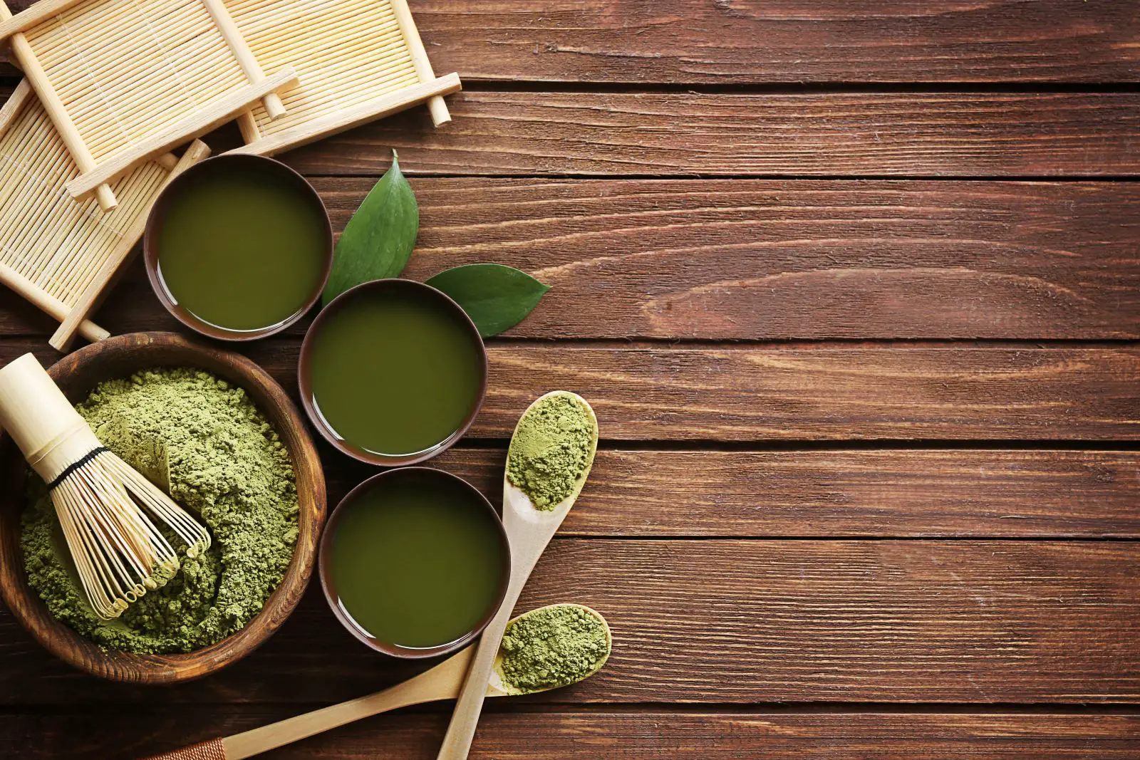 three small cups of green tea and a bowl and two spoons with matcha powder on a wooden tabletop
