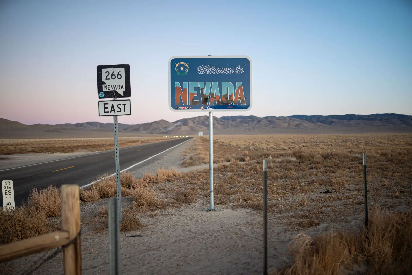 Top 5 Places to Retire in Nevada- Highway 266 East sign along the highway and a sign that says Welcome to Nevada on the right side, a barbed wire fence with metal poles on the lower part of the photo, desert on both sides and brown foothills in the distance