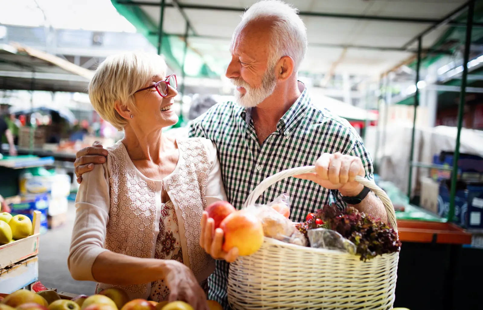 Grocery Store Discounts for Seniors- A senior couple smiling at each other holding a wicker basket of vegetables in a grocery store 