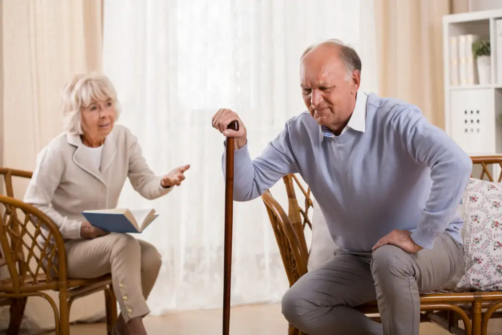 A senior woman encourages a senior man to use a walker. 