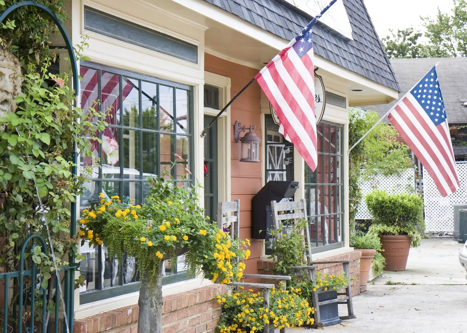 Small town street showing store fronts with green plants, some with yellow flowers on the sidewalk and flags on the corner of the buildign and by the door of the store.