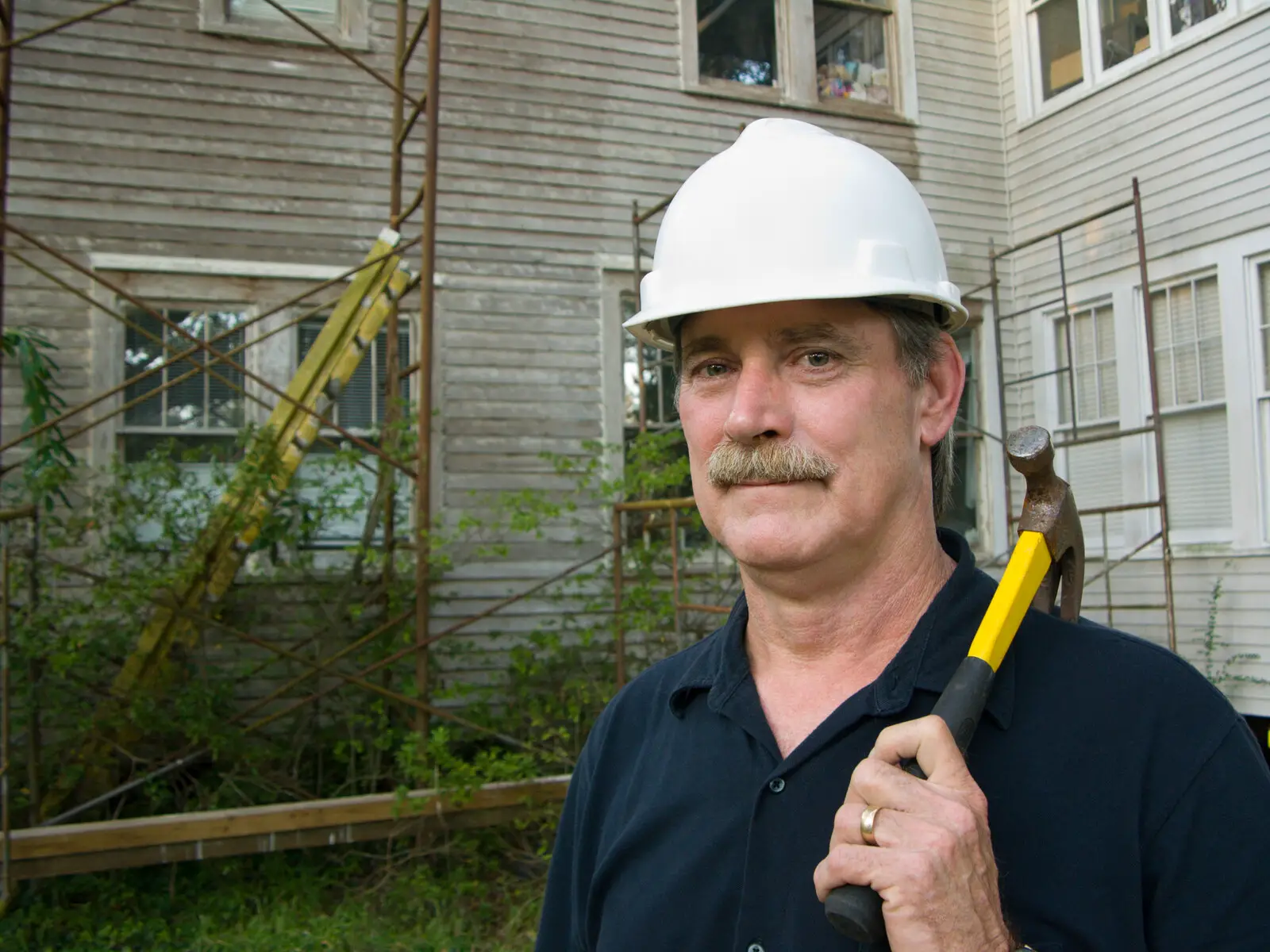 Home Repair Scams Targeting Seniors - Man holding a hammer restring on his shoulder with a house in the background with scaffolding and overgrown shrubs around it