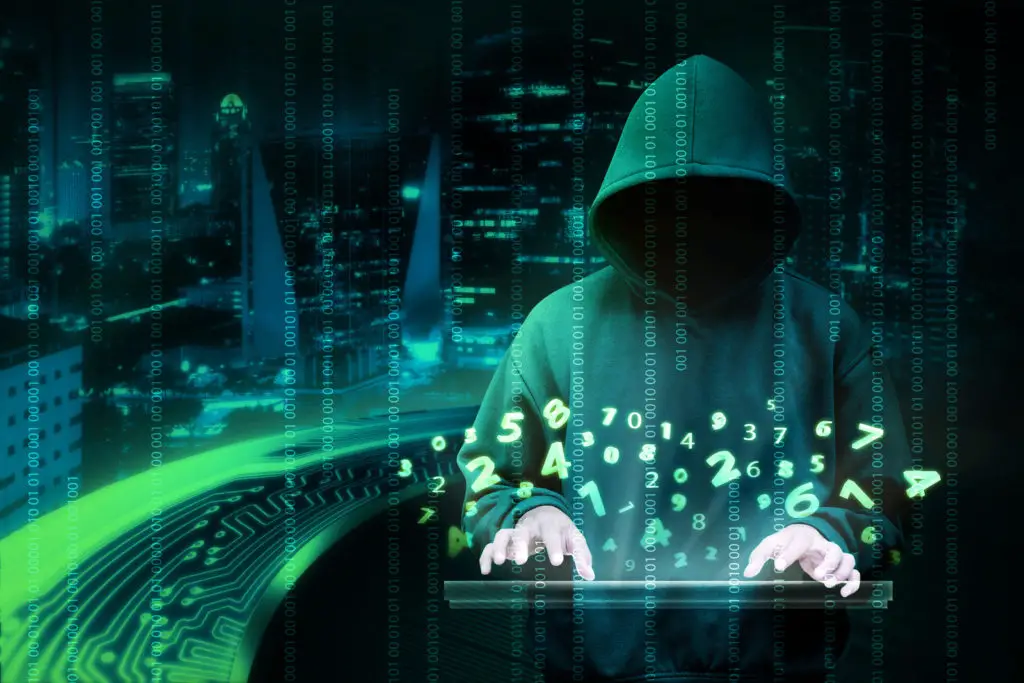 Dark background of a cityscape at night with a  Man in hoodie typing on a computer keybpard with a hologram of numbers and random data overlaid on the screen-represents a cyber criminal hacking into a database