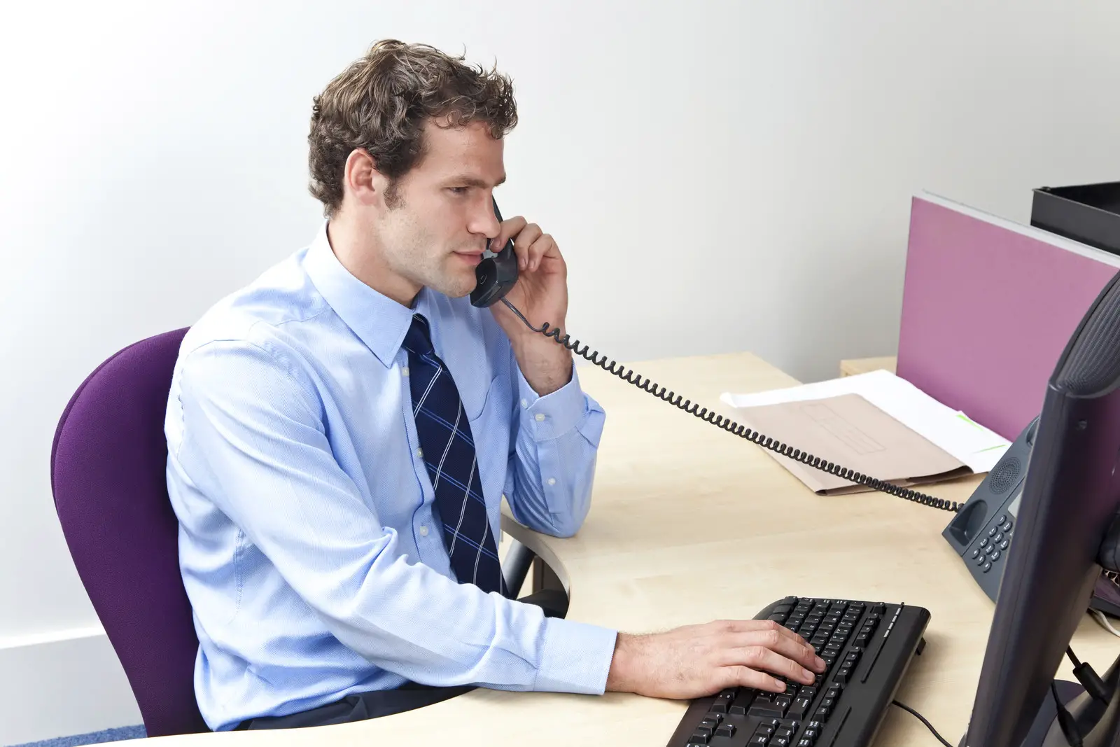 Salesman sitting at desk on the phone typing on the computer-represents an insurance agent attempting to sell upgraded life insurance policies