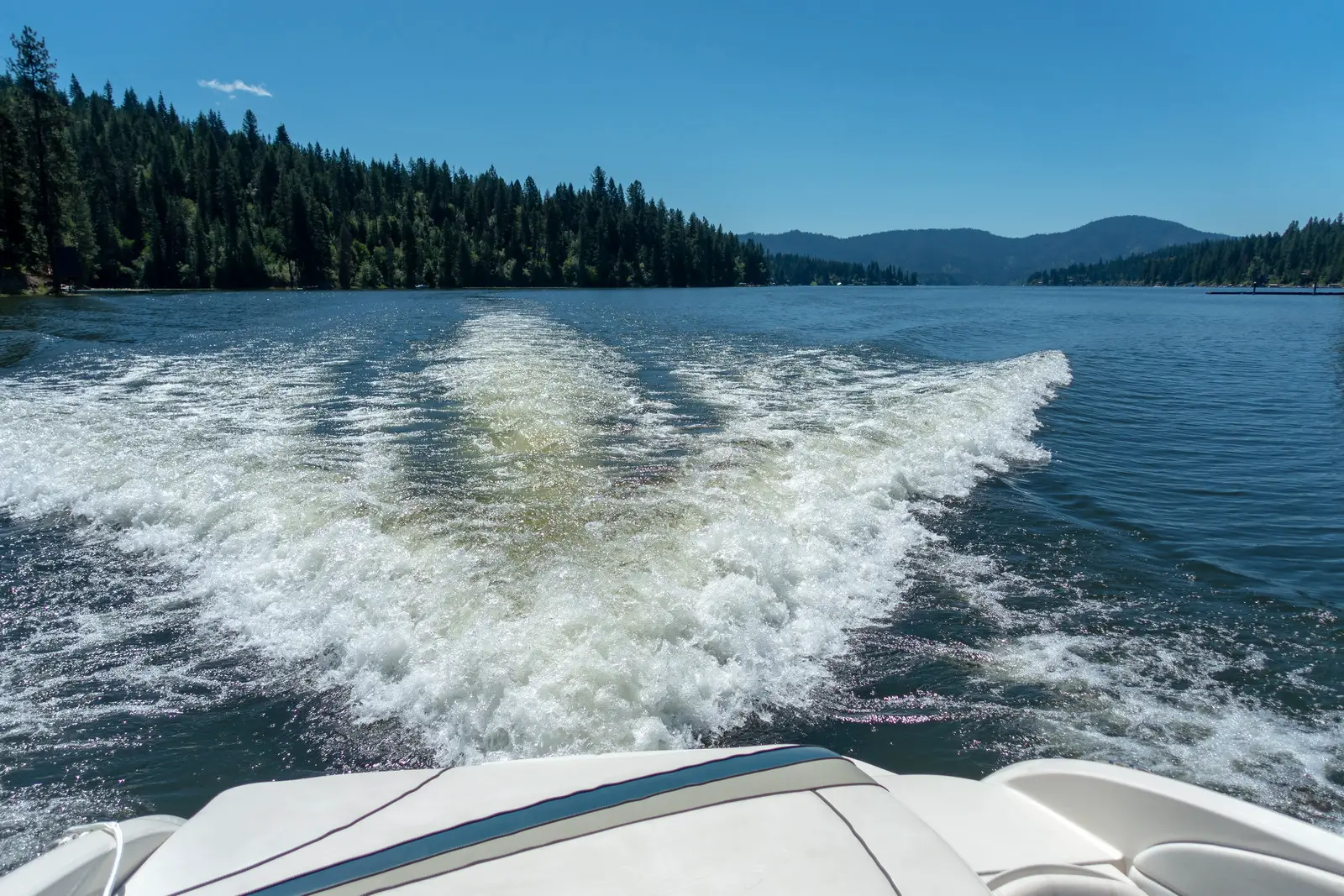Photo from the back of a boat on Hayden Lake with green pines lining the lake and mountains in the background