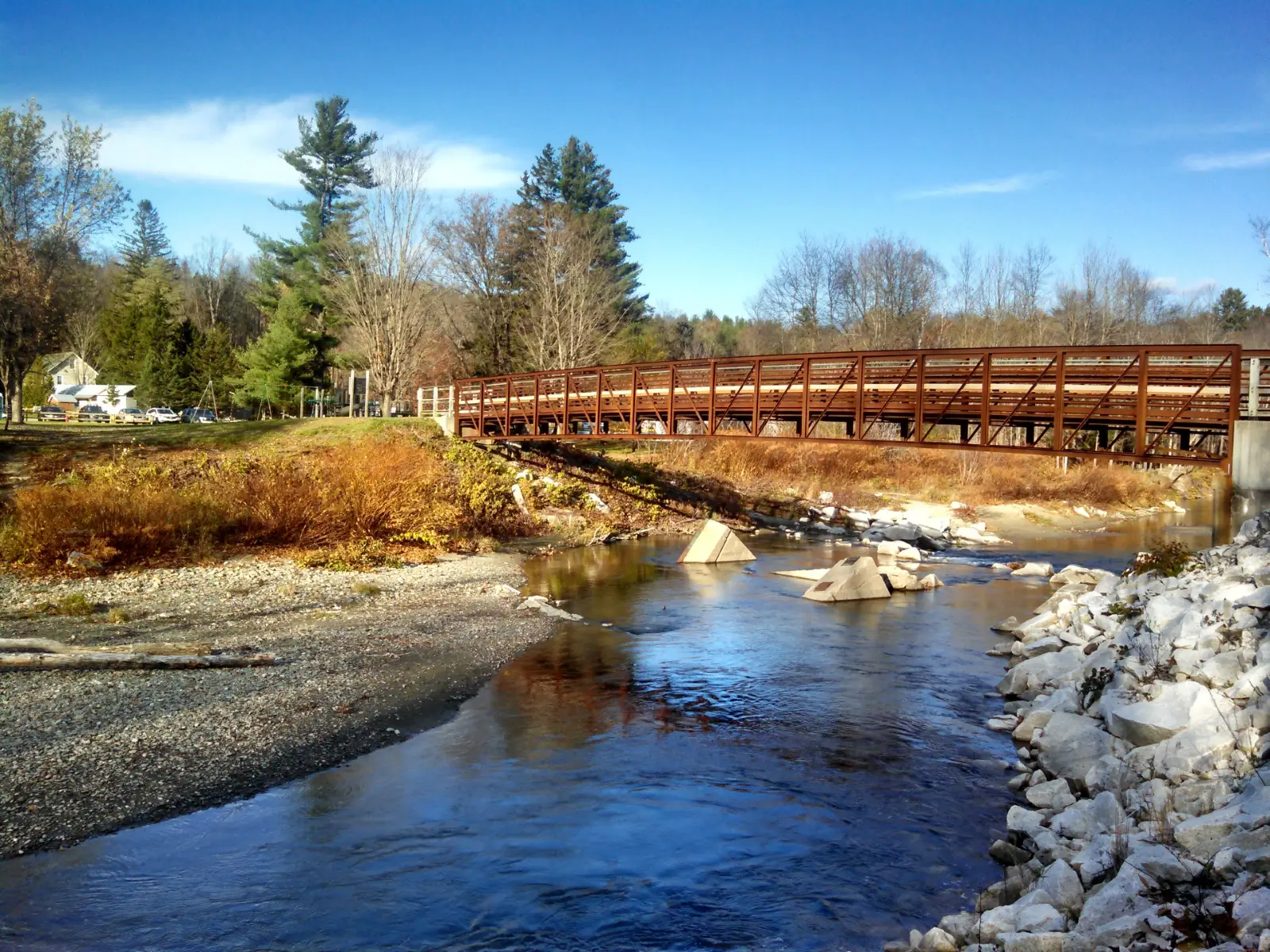 Fall day with blue skies and a brown pedestrian bridge over a river with a rocky riverbank - a small farmhouse in the background with pines and trees in the background