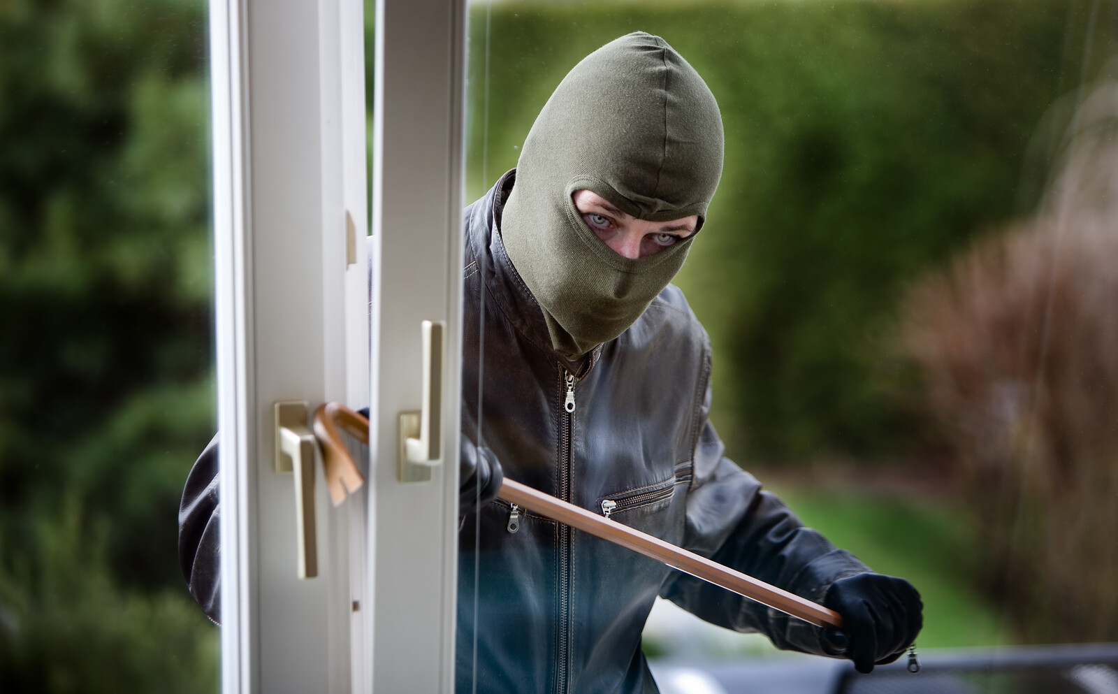 A burglar breaking into a house with a crow bar  through a window, the burglar has a green mask, leather coat, and black gloves 