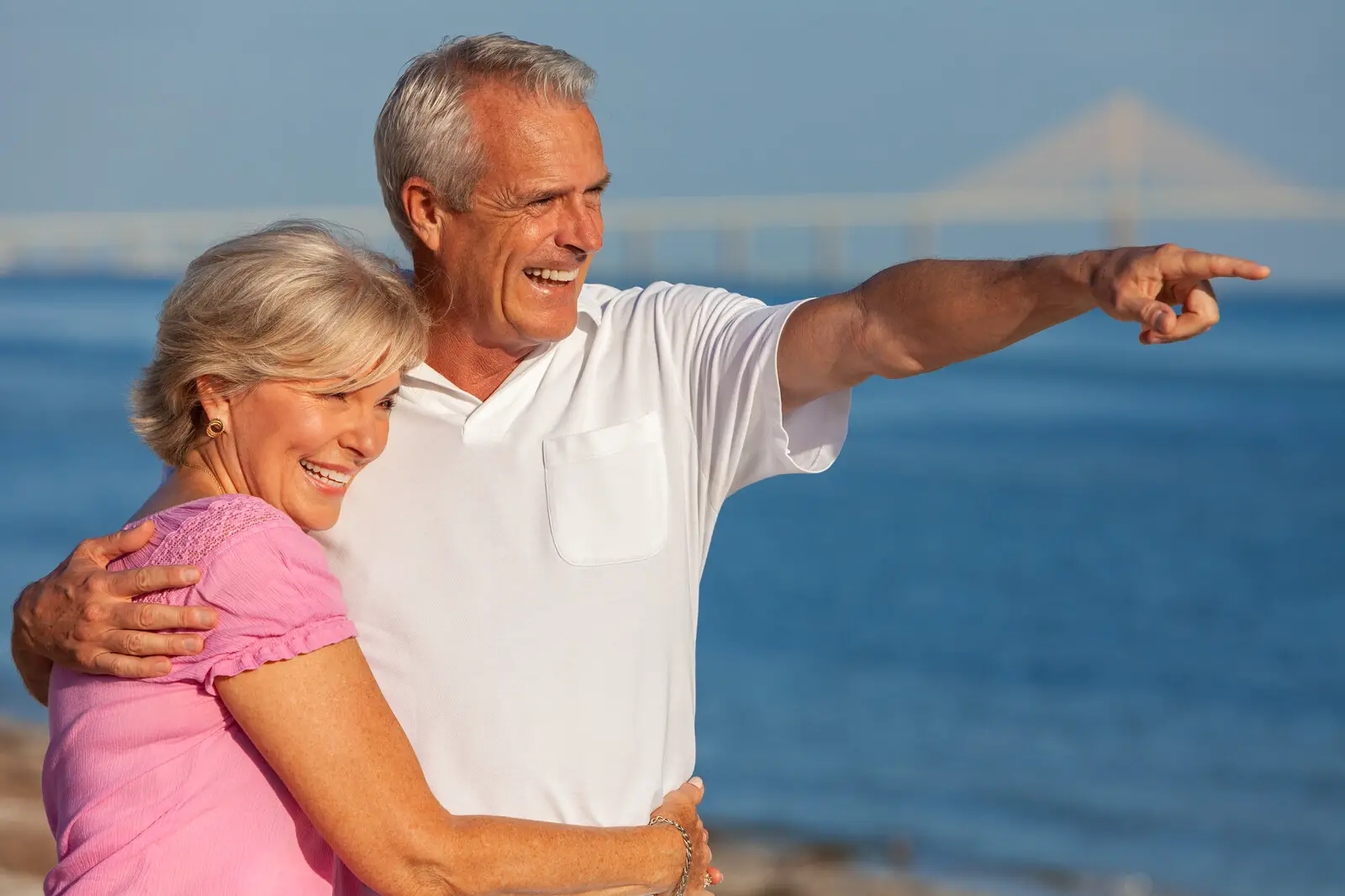 Travel Scams  - A happy smiling senior couple standing on a beach embracing the man is pointing out further along the ocean behind blue skies and a faded bridge in the background. 