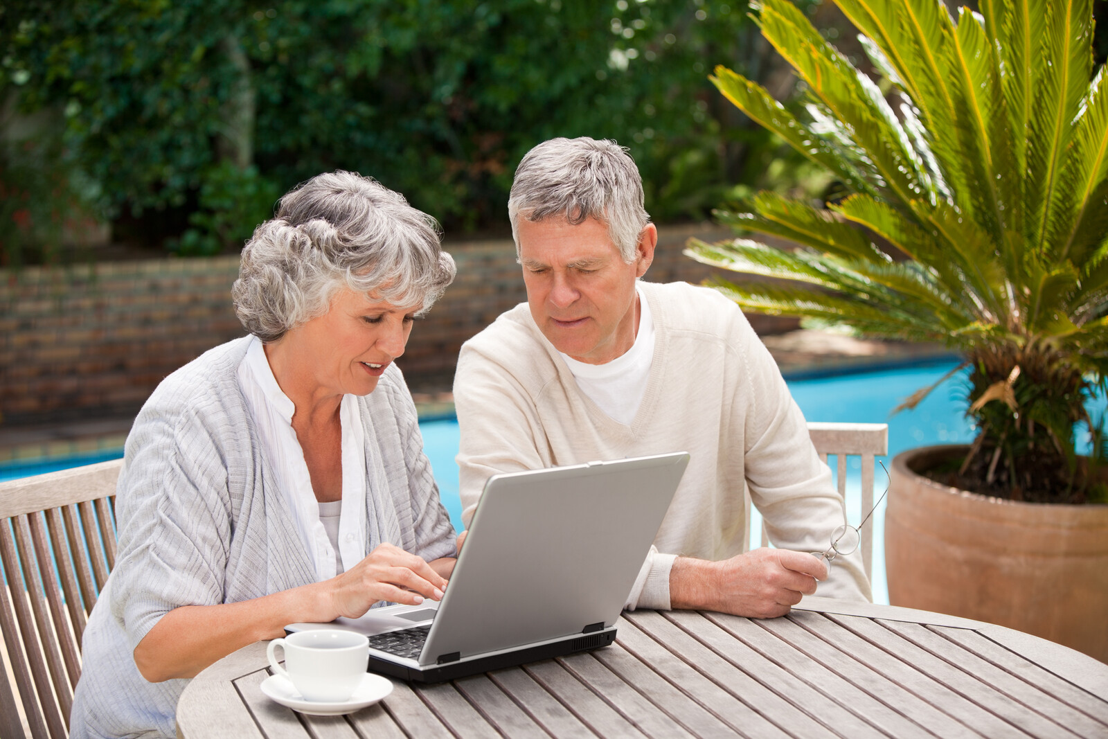Senior couple looking at a computer while sitting outside at a wood picnic table near a pool - represents seniors ordering vacation certificate